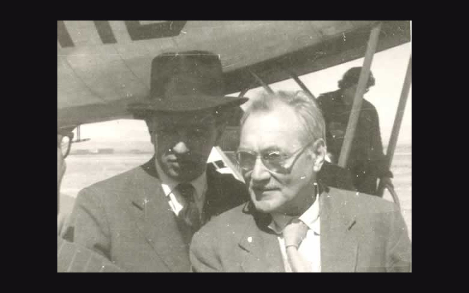 Two men stand near an aircraft, one in a hat and suit, the other bespectacled