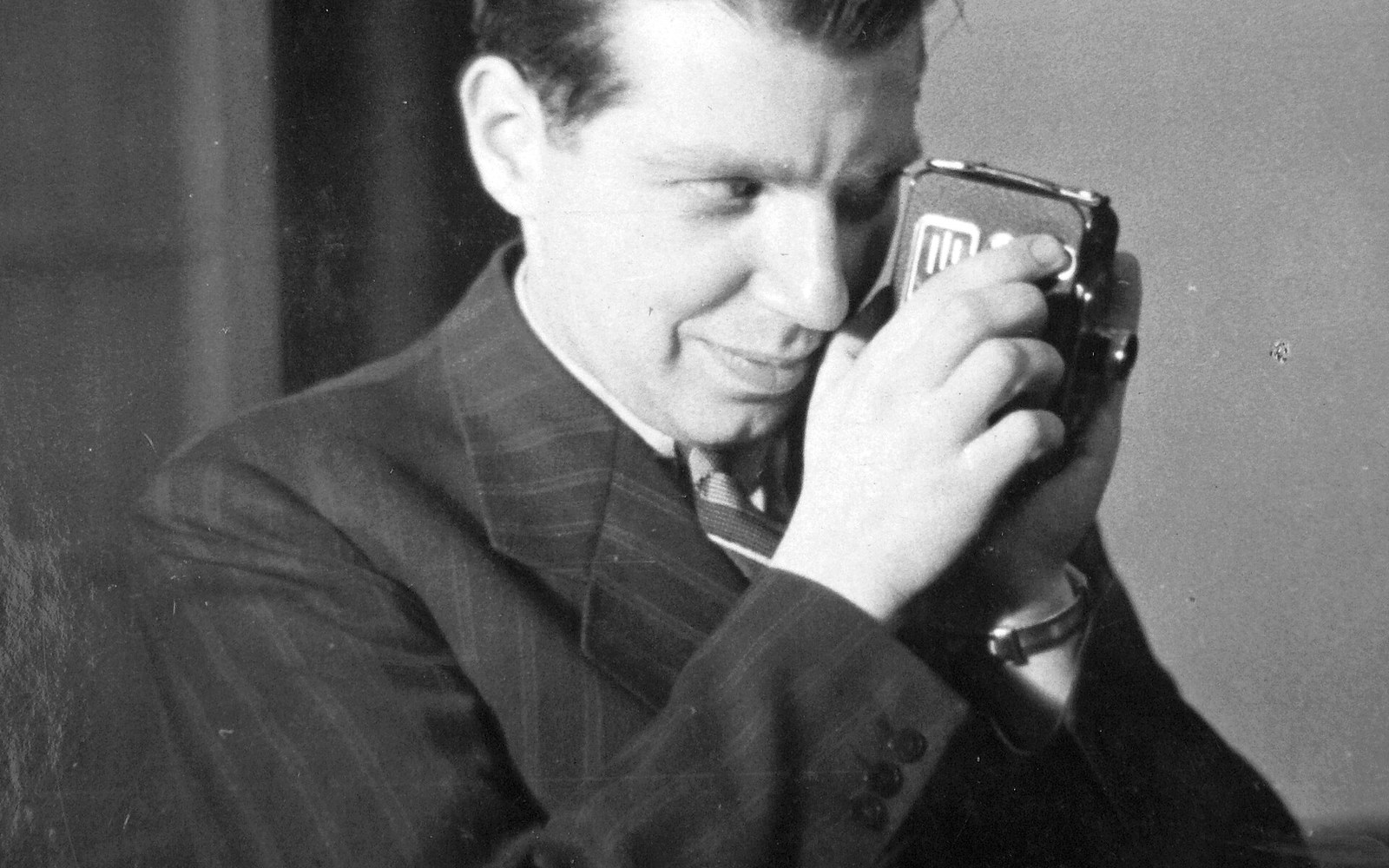 A man in a pinstriped suit holds a small camera up to his eye